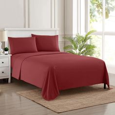 a bed with red sheets and pillows in a room next to a white dresser, potted plant and window