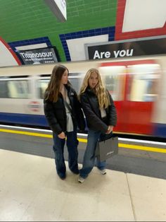 two young women standing in front of a train at a subway station while the train passes by