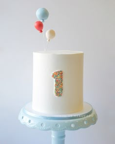 a white cake with sprinkles and balloons on top is sitting on a blue stand