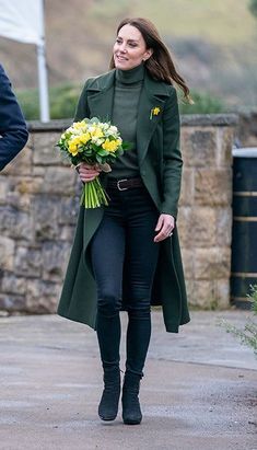 the woman is walking down the street with flowers in her hand and she is wearing a green coat