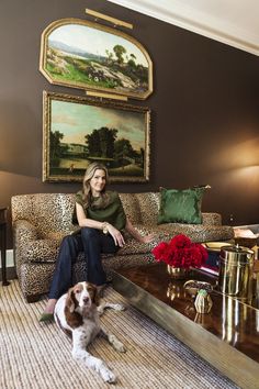 a woman sitting on top of a couch next to a brown and white dog in front of a painting