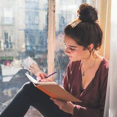 a woman sitting on a window sill writing with a pen and paper in her hand