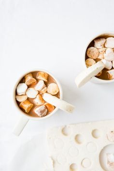 two mugs filled with marshmallows on top of a white table next to a piece of cheese