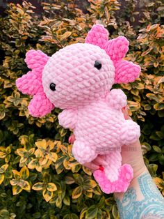 a hand holding a pink crocheted stuffed animal in front of some shrubbery