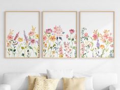 three floral paintings hang on the wall above a white couch in a living room with pillows