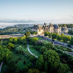 an aerial view of a castle surrounded by trees and fog in the distance, with mountains in the background