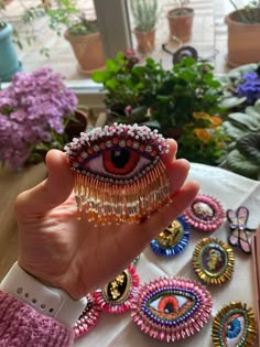 a person holding up an eye brooch in front of some potted plants and flowers