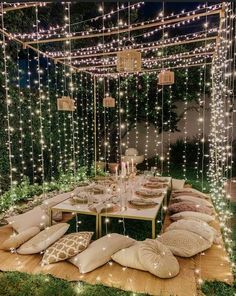 an outdoor dining area with lights and pillows on the ground, surrounded by greenery