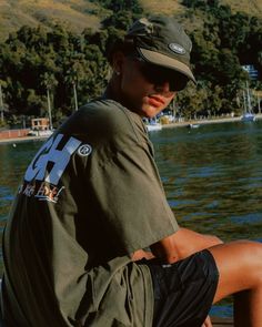 a young man sitting on the edge of a lake wearing a hat and green shirt