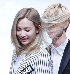 two people standing next to each other in front of a white wall and one person with blonde hair