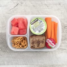 a plastic container filled with food on top of a wooden table next to carrots and pretzels