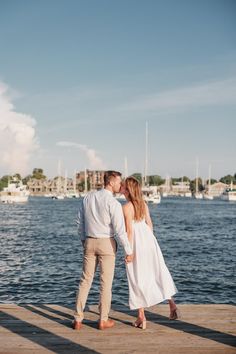 a man and woman standing on a dock next to the water with boats in the background