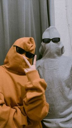 two people dressed in hoodies and sunglasses sitting next to each other, one holding the peace sign