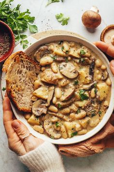 two hands holding a bowl of mushroom soup with bread and parsley on the side