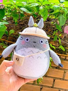 a hand holding up a small crocheted toy in front of some plants and flowers