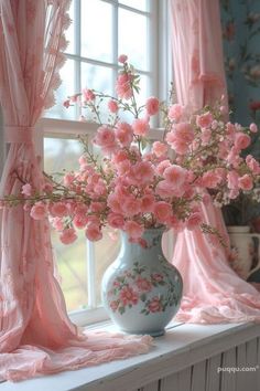 a vase with pink flowers sitting on a window sill in front of a curtained window