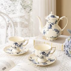 an open book on a table next to two tea cups and saucers with blue flowers