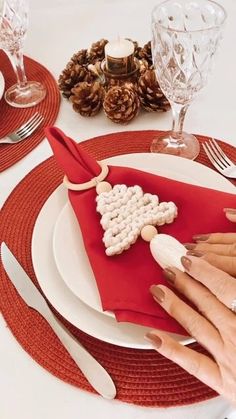 two hands are decorating a red napkin on a white plate with silverware and pine cones