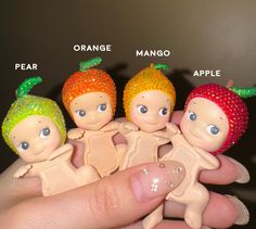 three little dolls are sitting in the palm of someone's hand, with oranges and mangos on them