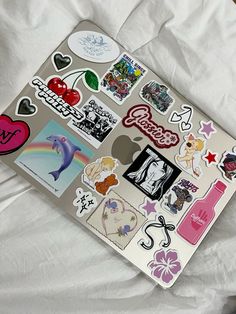 an apple laptop covered in stickers on top of a white sheet with sheets and pillows