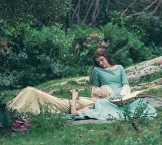 two people sitting on the ground reading books in the grass with trees and bushes behind them