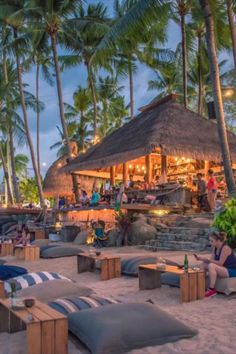 people are sitting on the beach at night with palm trees and lounge chairs in the foreground