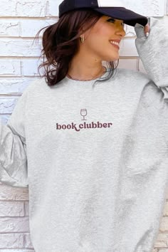 a woman wearing a book clubber sweatshirt standing against a brick wall with her hands behind her head