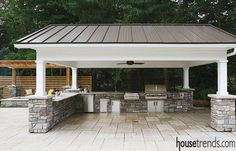 an outdoor kitchen and grill area in a backyard with stone walls, white pillars, and brick pavers flooring