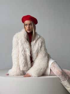 a woman sitting on top of a white table wearing a red hat and fur coat