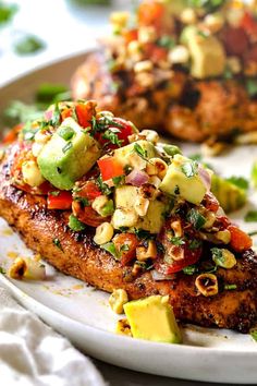 two pieces of meat on a white plate with avocado and tomato toppings