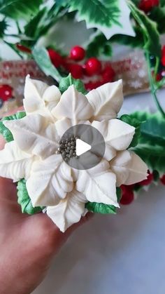 a hand holding a white flower with green leaves and berries around it on a table