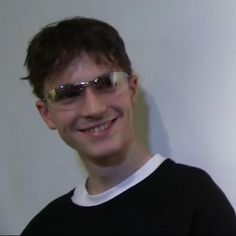 a young man wearing sunglasses and smiling for the camera while standing in front of a white wall