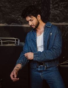 a man wearing a denim jacket and jeans standing in front of a black wall with his hand on his hip