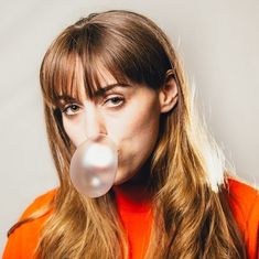 a woman with long hair blowing a bubble in front of her face while wearing an orange shirt