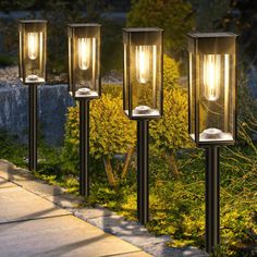 three outdoor lights are lit up on the sidewalk in front of some bushes and trees