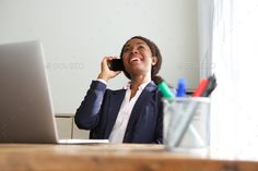 a woman sitting in front of a laptop talking on her cell phone and smiling at the camera