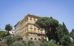 an old building is surrounded by trees and people walking on the sidewalk in front of it