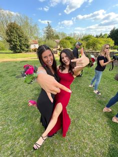 two women in dresses giving the thumbs up sign while standing on grass with other people behind them