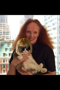 a woman holding a cat in front of a large window with city buildings behind her