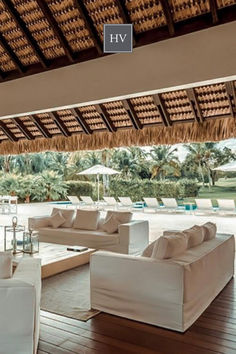 a living room filled with white couches under a thatched roof next to a swimming pool