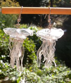 two clear glass jellyfish earrings hanging from a wooden rod in front of some bushes