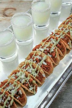 several tacos are lined up on a tray with drinks in glasses behind them,