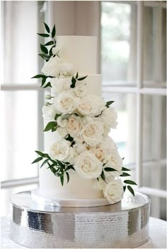 a wedding cake decorated with white flowers on top of a silver platter in front of a window