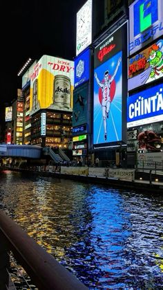 there are many billboards along the water in this city at night, with lights reflecting on the water