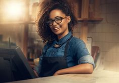 a woman wearing glasses and an apron smiles while looking at the camera with a laptop in front of her