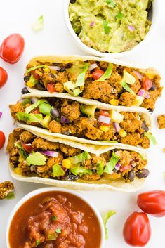 three tacos with guacamole, tomatoes and lettuce on the side