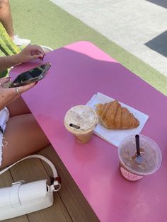 a woman sitting at a pink table holding a cell phone and eating croissants