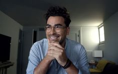 a man with glasses is smiling and looking at the camera while sitting in his living room