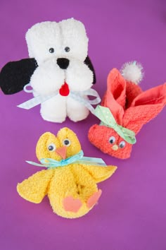 three small stuffed animals sitting next to each other on a purple surface with one yellow duck