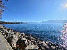 the sun shines brightly over water and rocks on the side of a road next to a body of water with mountains in the distance